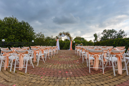 Open air decorated area for the wedding ceremony with a wooden arch decorated with flowers. White wooden chairs decorated with orange bows and roses outdoors on green grass background.の写真素材