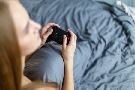 Girl gamer playing video game with wireless joystick at home. Gamepad in female hands close-up, gaming addiction concept, woman gamer in underwear relaxes in the bedの写真素材