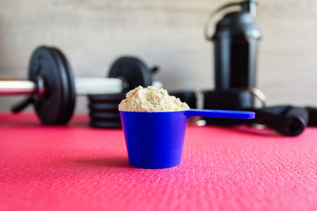 Plastic spoon or measuring scoop of whey protein on the background of sports equipment, a shaker for mixing the mixture, black dumbbells, leg ties, elastic bands for traction and attachment handles.の写真素材