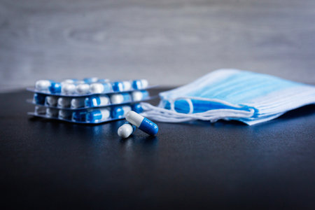Blue-and-white capsules with drug on background of disposable medical protective masks cover the mouth and nose, as well as stacks of Packed capsules. Healthcare and medical concept of covid-19の写真素材