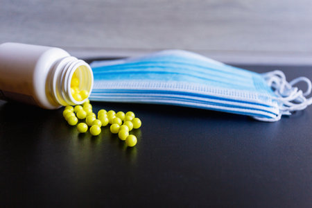 Scattered yellowish round tablets, vitamins scattered from a white plastic bottle, and a blue protective medical face mask on a black background. Prevention of covind-19 or coronavirus. Close upの写真素材