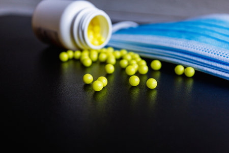 Scattered yellowish round tablets, vitamins scattered from a white plastic bottle, and a blue protective medical face mask on a black background. Prevention of covind-19 or coronavirus. Close upの写真素材