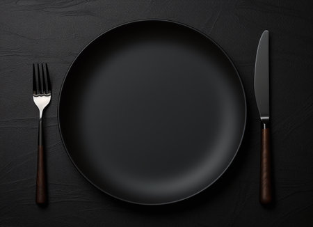Empty black plate with knife and fork on black background, top viewの素材