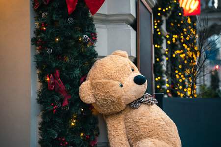 Big teddy bear with a bow tie sits by Christmas decorations. Lights and garlands make the street...