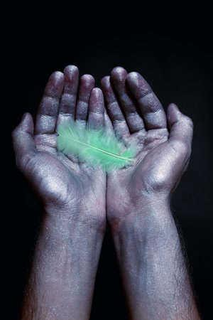 Strong male hands in silver paint holding light green feather on  black backgroundの写真素材
