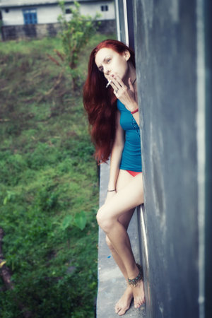 Beautiful young woman with long red hair, smoking in the windowの写真素材