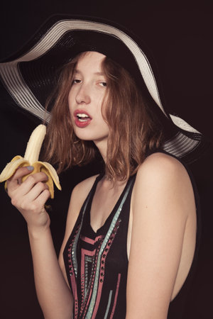 Girl dressed in summer hat eats a banana. Studio photo on  black backgroundの写真素材