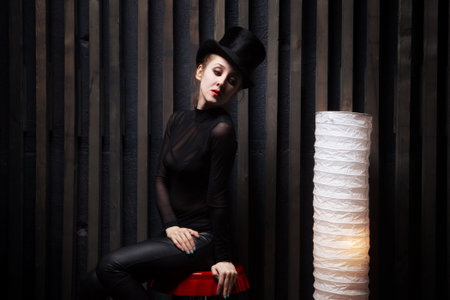 Beautiful young woman wearing  top hat Sits on  chair near to a floor lamp in a dark roomの写真素材
