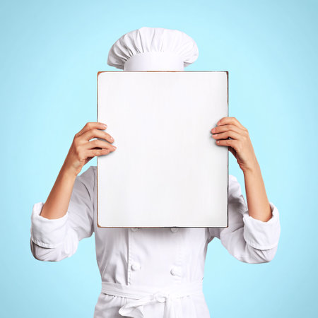 Young female chef hiding her face behind blank paper sheet on blue backgroundの素材