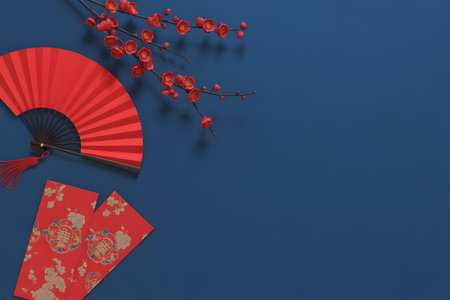 Chinese new year items traditional red folding fan, lucky red envelopes, and cherry blossom branches on a deep blue backgroundの素材