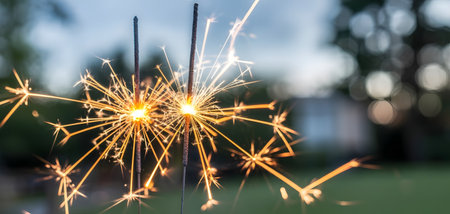 Closeup of two sparklers burning brightly with golden sparks against a soft, blurred evening background, perfect for celebrationsの素材