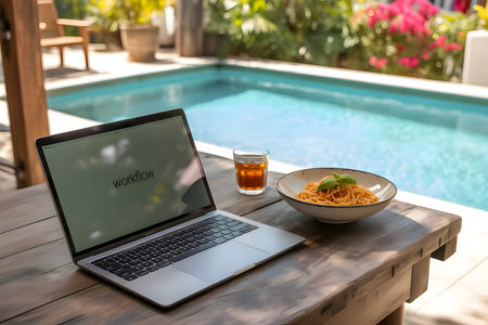 Laptop with 'workflow' on screen, a fresh spaghetti meal and a drink on a wooden table beside a resort swimming pool, representing remote working.の素材