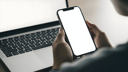 Person using a smartphone and laptop in a modern workspace with a blurred backgroundの素材