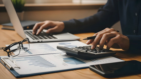 Business professional calculating finances and working on a laptop at a wooden desk indoorsの素材