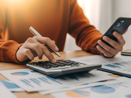 Person calculating finances with a calculator and smartphone at a bright deskの素材