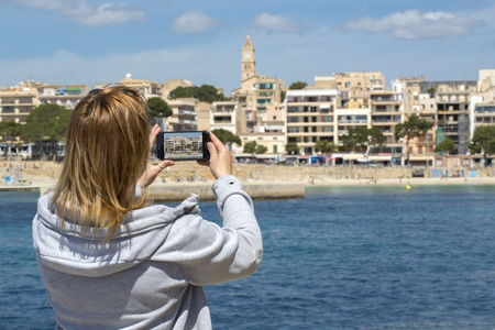 Young blonde woman sightseeing and taking photos with her smartphone in Porto Cristo, Spain.
