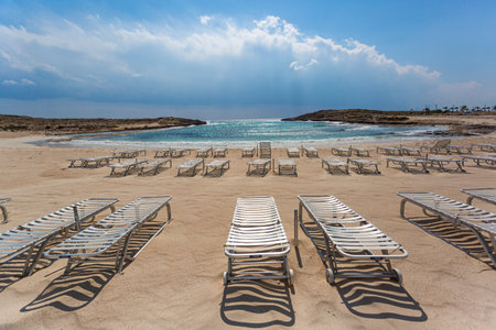 Rows of empty sunbeds stand on a sandy beach, inviting tourists to relax by the clear turquoise waterの写真素材