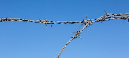 Barbed wire fence forming ay shape against a clear blue sky, symbolizing division, barrier, or...