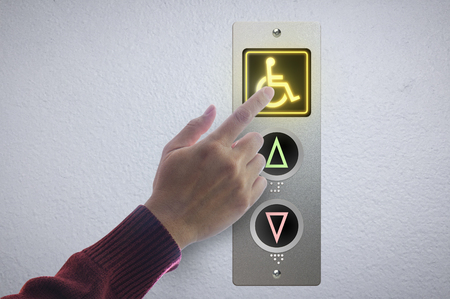 A hand pressing a button designed especially for handicap on an elevator button panel with an amber glowing lightの写真素材