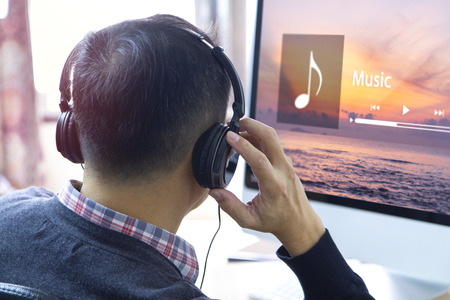 Back side of an Asian man wearing black heaphones listen to internet music streaming with relax and calm in front of computer screen while taking a break during routine work.の写真素材