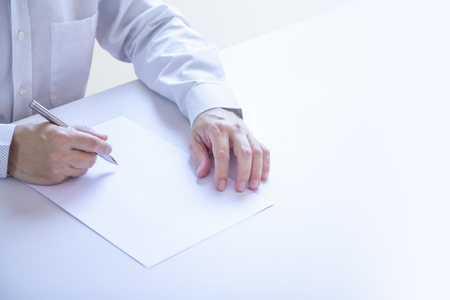 Business man partially cropped at his hands holding a pen with writing gesture on a plain blank white paper document as a blank mock-up template to be refilled with any information.の写真素材