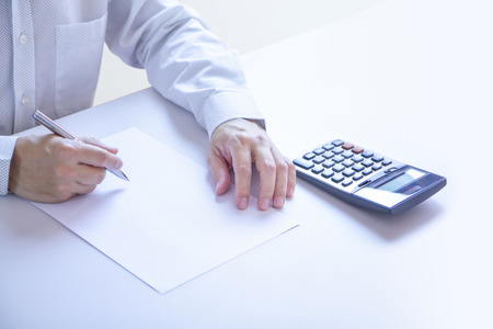 Business man partially cropped at his hands holding a pen in writing gesture on a plain blank white paper document with a calculator as a blank mock-up template to be refilled with any information.の写真素材