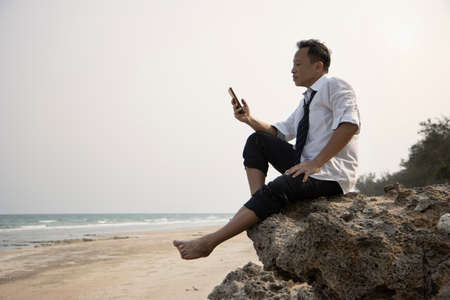 Relax Asian businessman in casual cloth sitting on a cliff at beach with less tense or anxious at...