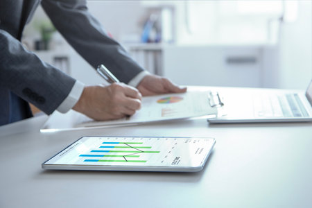 Businessman standing in the office reviewing financial annual reports with modern tablet and laptop computer for a return on investment.の写真素材