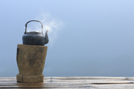old and black retro kettle for coffee or tea with hot steam or smoke place on wood platform and table or floor in the morning at countryside with sea of mist or fog background and copyspaceの写真素材