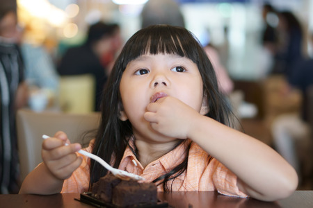 Asian children cute or kid girl enjoy and fun with happy eating delicious brownie chocolate cake and took hand in mouth for sweet dessert on wood table and holding spoon at lunch in cafe restaurantの写真素材