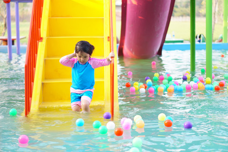 Asian children cute or kid girl on swimming pool with colorful ball on blue water and smile with happy fun on stairway and slider in water park for refreshing and relax with exercise on summer holidayの写真素材