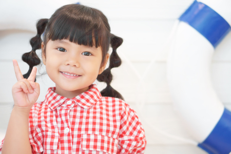 Asian children cute or knit braid kid girl wear a red plaid shirt on the boat with blue life ring and two finger show for confident and white teeth smile on soft focus with spaceの写真素材