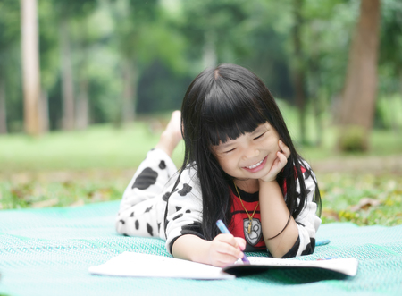Asian children cute or kid girl learning for coloring and sleep prop up or hand to cheek with drawing paint on green mat and nature meadow at garden with smile white teeth for fun on holiday relaxの写真素材
