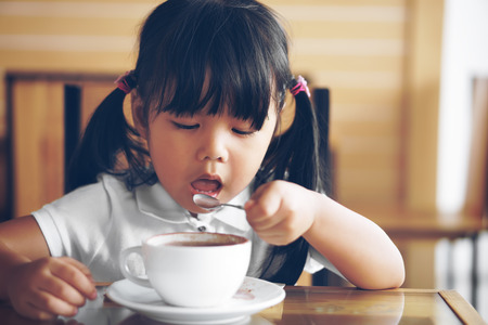 Asian children cute or kid girl holding spoon and open mouth for delicious hot cocoa or chocolate drinking in white cup for breakfast in the morning on table in home or cafe restaurant with vintageの写真素材