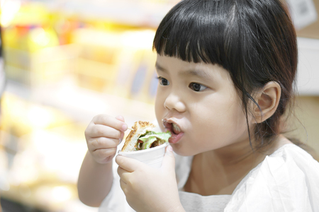 Asian children cute or kid girl taste and eating sandwich or hamburger for delicious or not delicious food in the supermarket at department storeの写真素材