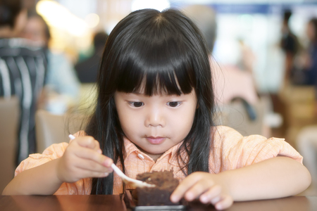 Asian children cute or kid girl enjoy and fun with happy eating delicious brownie chocolate cake for sweet dessert or snack on wood table and make eyebalsl at lunch in restaurant or cafeの写真素材