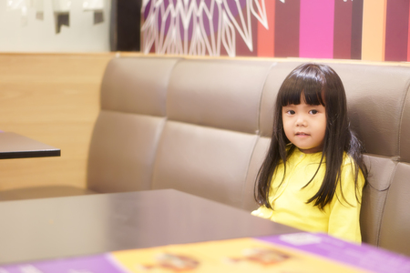 Asian children cute or kid girl relax sitting for waiting order or food for lunch or dinner on long sofa and wood table at cafe restaurant with wear yellow shirt and copy spaceの写真素材