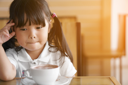Asian children cute or kid girl has dimple and hot cocoa or chocolate drinking in white cup for breakfast with thinking or absent-minded and strain in morning on table in home or cafe with sunlightの写真素材
