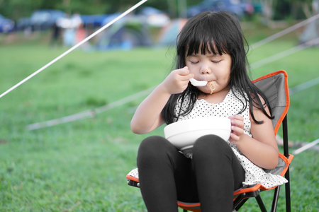 Asian children cute or kid girl and disheveled hair with hungry eating instant noodle delicious and sit on camping chair with green grass at nature camping tent trip on summer or rainy season holidayの写真素材