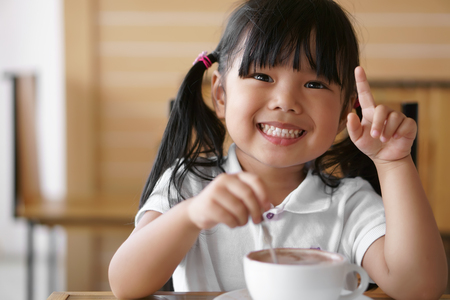 Asian children cute or kid girl white teeth smile and hot cocoa or chocolate drinking for breakfast and want one more because delicious in the morning on wood table in the home or cafeの写真素材