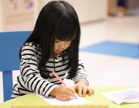 Asian children cute or kid girl learning for coloring or hand drawing paint on white paper and colorful table with chair at nursery or pre school on soft focusの写真素材