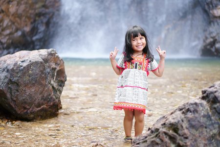 Asian children cute or kid girl smile with fun and enjoy playing with show two finger for take a photo on waterfall with stone or rock and holiday relax trip for summer at Jokkradin waterfallの写真素材