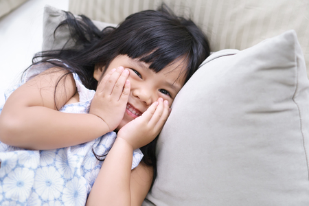 closeup Asian children cute or kid girl wake up in morning or sleeping and open eye with white teeth smile and hand take to cheeks or shy on white bed and bedtime in bedroom at home or hotelの写真素材