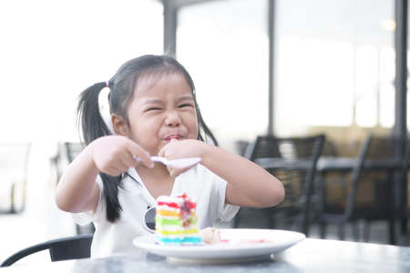 Asian child cute or kid girl smiling enjoy and fun with happy eating delicious rainbow cake and suck finger for sweet and sour dessert on table and holding spoon at lunch in cafe restaurant or shopの写真素材