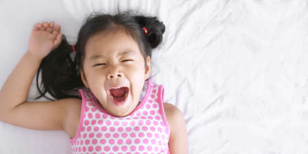 Asian child cute or kid girl sleep yawn and open mouth with stretch oneself after wake up late and sleepy in morning on white bed in bedroom for relax on holiday and wear pink dress with copy spaceの写真素材