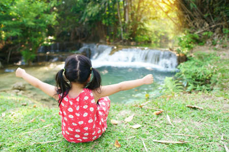 Asian child or kid girl sit and happy hands up on nature clear waterfall with green grass and tree in jungle or forest for holiday relax or camping picnic and summer travel trip on family vacationの写真素材