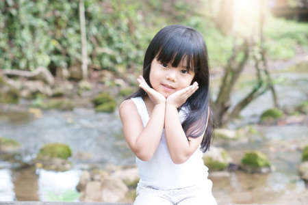 Asian child cute or kid girl cheerful and smile with hand to cheek on face and nature stream in jungle or green garden and public park with tree for beauty fashion on summer holiday relax and happyの写真素材
