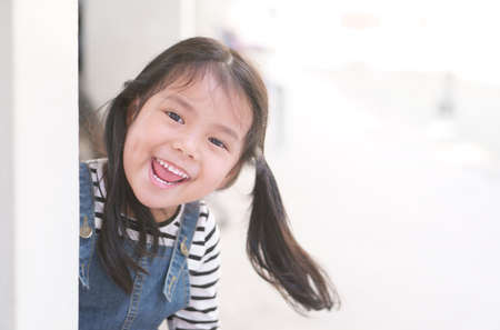 Asian child cute or kid girl playing peekaboo or hide and seek with sneaked or dodge behind the post and happy smiling white teeth or cheerful at public park or playground on relax holiday with spaceの写真素材