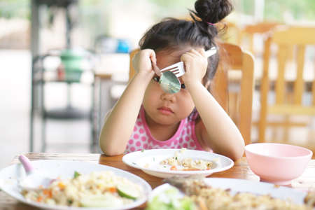 Asian child or kid girl anorexia or sad and bored food or boredom with sleepy vacant and prop up or hand to head with headache on wood table for breakfast or lunch eating at restaurant on preschoolの写真素材