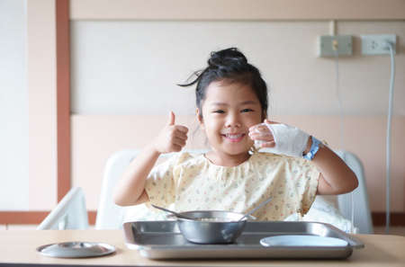 Asian child or kid girl Influenza A sick with happy smiling and show thumb to good encouragement with salt water in hand blood vessels on bed at hospital room for treatment healthcare with lunch foodの写真素材